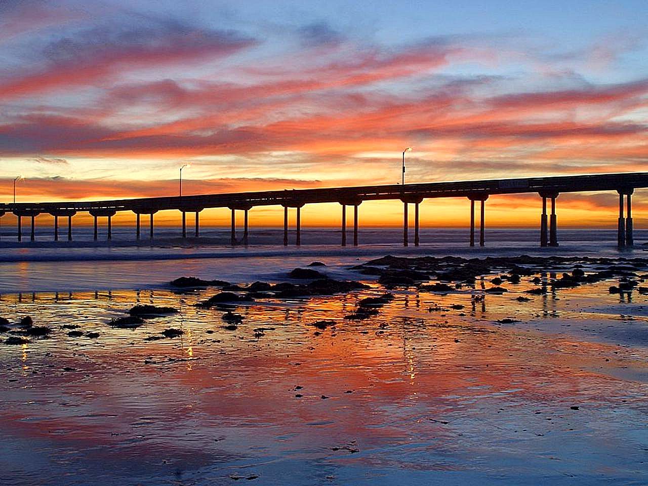 The Ocean Beach Pier at sunset, a long concrete pier on pilings over wet sand and rocks, with an orange sky.